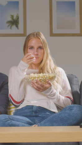 Vertical-Video-Of-Or-Couple-Friends-Sitting-On-Sofa-At-Home-Watching-TV-And-Eating-Popcorn-